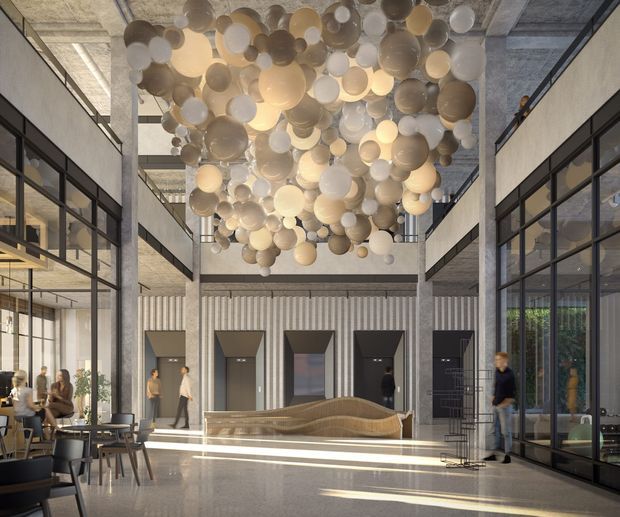 A spacious contemporary commercial lobby or atrium with exposed concrete ceilings and columns, extensive black-framed glass partitions, a polished light floor, elevator doors, and a sculptural curved reception desk. A large suspended installation of translucent spherical forms creates a dramatic feature above the central space, while café-style seating and indoor planting occupy the sides.