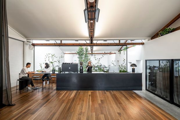 Open-plan contemporary office reception and workspace with a long matte-black reception counter, timber flooring, exposed timber-and-steel ceiling structure, large glazed meeting-room partitions, and a bright planted courtyard visible beyond. A few people work at a small table to the left.