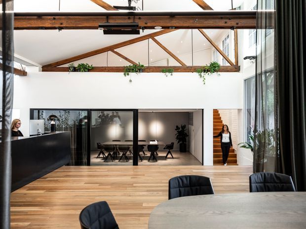 A bright, double-height commercial office or studio interior with exposed timber beams and triangular roof trusses, white walls, warm timber flooring, black-framed glass meeting rooms, a timber stair, indoor planting, and a large communal meeting table in the foreground. The space has a contemporary adaptive-reuse feel with visible structural timber and abundant glazing.