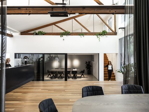 A bright, open-plan commercial interior with a large communal meeting or dining table in the foreground, black upholstered chairs, a reception counter at left, and a glazed meeting room beyond. Exposed dark timber beams and trusses span the high ceiling, with hanging greenery, extensive glazing, timber flooring, black-framed partitions, and a visible timber stair. The space has a refined contemporary workplace or hospitality character.