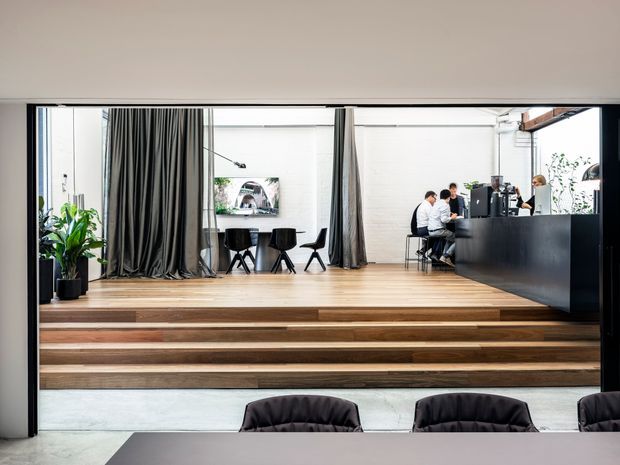 Contemporary commercial hospitality or café interior viewed from a lower seating area. A raised timber platform with broad stepped risers leads to a counter and coffee service area, with a small dining setting, dark curtains, indoor plants, exposed white-painted brick walls, and black architectural framing. The foreground contains a large communal table and upholstered seating.