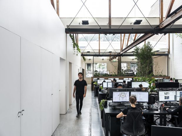 A bright, open-plan commercial office in a converted industrial-style space. Rows of black workstations with computer monitors occupy the right side, while a clear circulation path runs alongside tall white built-in storage or partition walls on the left. The space has a polished concrete floor, exposed dark structural beams, extensive overhead glazing, timber framing, hanging greenery, and large indoor plants. Several people are working at desks or walking through the office.