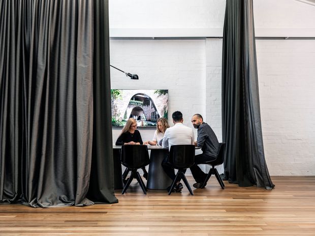 A bright, high-ceilinged commercial interior with white-painted brick walls, warm timber flooring, and dramatic full-height dark curtains framing a small meeting area. Four people sit around a round table on black chairs, with an artwork and minimal wall lighting in the background.