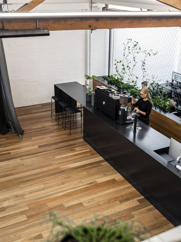 A high-angle view of a contemporary commercial café or coffee bar interior. A long matte-black service counter with espresso machines and coffee equipment runs along the right side, with a barista working behind it. Black bar stools and a narrow dining counter sit against a white-painted brick wall. Warm timber flooring and exposed timber beams contrast with the black joinery, while planted greenery lines the glazed window wall. The space has an industrial, loft-like character with abundant natural light.
