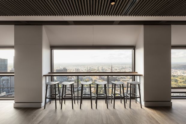 A refined high-rise interior with a long slim timber-look counter and six bar stools positioned in front of expansive city-facing glazing. Dark vertical slatted ceiling detailing, pale structural columns, and timber-toned flooring create a contemporary hospitality or communal lounge setting, with an elevated urban outlook beyond the windows.