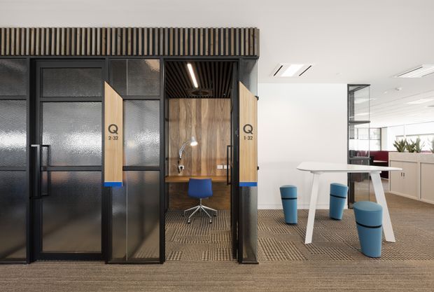 Contemporary commercial office interior featuring enclosed private work booths with black-framed textured glass doors, warm timber-panelled interiors, and integrated linear ceiling lighting. An adjacent open collaboration area includes a white high table, blue stools, carpeted flooring, and planter-lined glazing.
