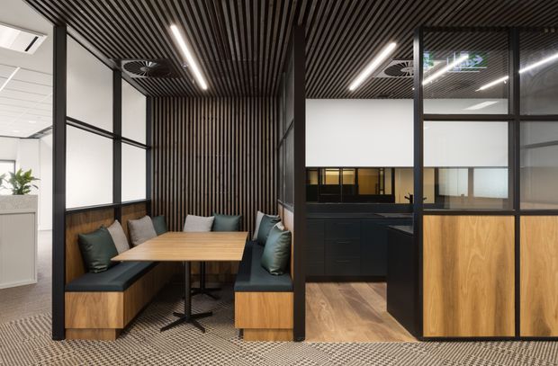 Contemporary commercial office breakout area with a built-in booth featuring timber seating bases, dark upholstered cushions, and a light timber table. Black-framed glazed partitions divide the booth from an adjacent kitchenette or workspace with dark cabinetry and timber panel fronts. The space has a dramatic black slatted ceiling with integrated linear lighting, patterned carpet, white walls, and black metal framing.