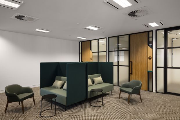 A contemporary office lounge and breakout area with dark green upholstered lounge seating arranged in semi-private high-backed booths, occasional black tables, and matching armchairs. The space has a neutral patterned carpet, white walls and ceiling, exposed rectangular and circular HVAC diffusers, recessed lighting, and black-framed glazed partitions with warm timber doors and panels.