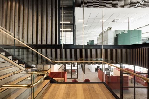 A contemporary multi-level commercial interior centered on a broad timber staircase and landing. Vertical timber slat wall and balustrade elements, black metal framing, clear glass partitions, brass-toned handrails, and warm timber flooring define the space. Open-plan offices with red upholstered seating are visible below, while a bright ceiling grid and glazed meeting rooms occupy the upper level.