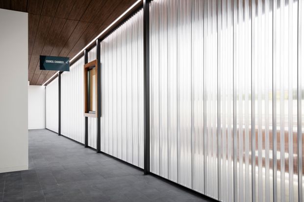 A contemporary commercial interior corridor with translucent vertically ribbed glazing forming meeting-room partitions on the right. The space has dark grey tiled flooring, a warm timber-lined ceiling, black-framed doors and mullions, and bright diffused daylight. A sign identifies a council chamber beyond the glazed partition.
