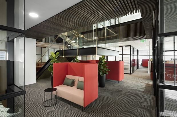 Contemporary commercial office interior with open-plan workspaces, red upholstered booth seating, black-framed glazed meeting rooms, indoor plants, a suspended mezzanine or stair structure, and a striking slatted timber ceiling. The space has a refined industrial character with layered circulation areas and patterned carpet flooring.