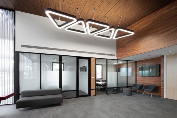 Contemporary commercial office reception or lounge area with a dark grey sofa and occasional chairs, framed glass meeting rooms, a wall-mounted display, and a striking geometric pendant light. The space combines grey tiled flooring, warm timber wall and ceiling lining, black-framed glazing, and a clean white partition wall.