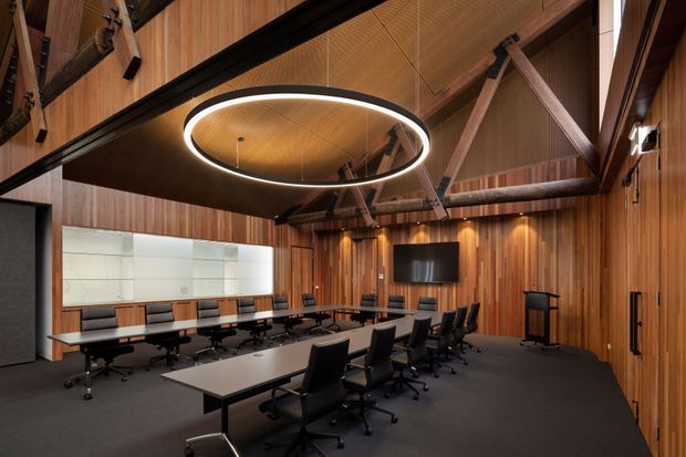 A contemporary boardroom or conference room with a long central meeting table surrounded by black office chairs. The room features extensive warm timber wall and ceiling panelling, exposed dark timber structural beams and braces, integrated recessed wall lighting, a large circular suspended LED light, dark carpet flooring, and a wall-mounted display. Glazed panels and a doorway are visible at the rear, creating a refined corporate interior.