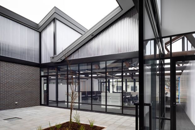 A contemporary internal courtyard or atrium within a commercial-style building, framed by dark brick walls, black metal-framed glazing, and angular roof forms. Translucent corrugated wall panels provide diffused light above the glazed office areas, while exposed structural beams are visible through the glass. The courtyard has pale tiled paving and a small landscaped planter, creating a bright, refined architectural setting.