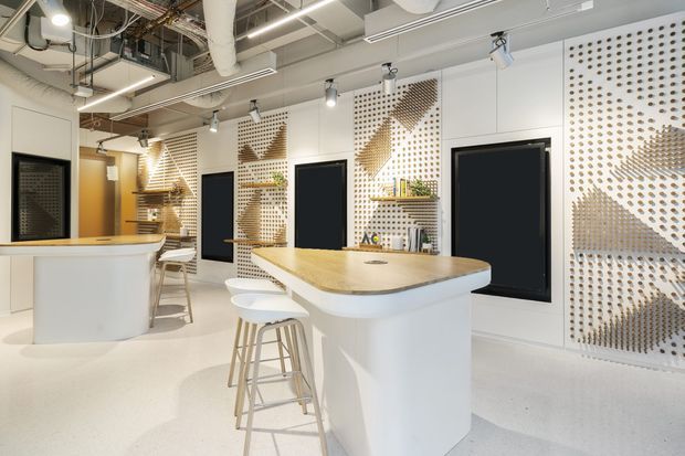 Bright contemporary commercial interior featuring white curved reception or service counters with pale timber tops, stools, black-framed openings, and decorative perforated white wall panels with geometric patterned inserts. The exposed ceiling services, track lighting, and open collaborative counter layout suggest a modern office or showroom fit-out.