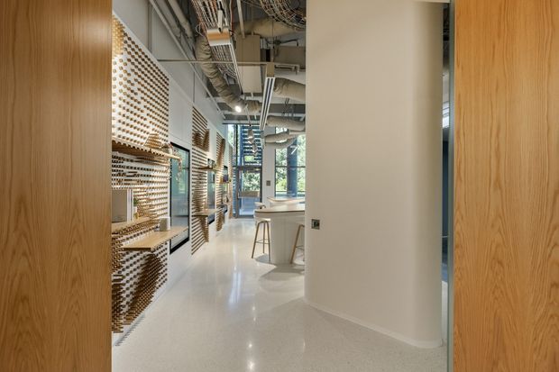 A bright contemporary commercial interior viewed through a timber-framed opening. The space has polished light terrazzo flooring, white curved columns and walls, exposed services and ductwork at the ceiling, and a linear café or kitchenette counter with stools. The left wall features extensive patterned timber or wood-toned display shelving and screens, while large windows bring in daylight at the rear.