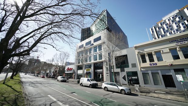 Street-level exterior view of a modern multi-storey commercial building in an urban streetscape. The central building has a glazed upper volume, a distinctive angular roof or facade form, and ground-floor retail or commercial tenancies. Adjacent buildings and mature street trees line the broad road, with parked cars and a marked cycle lane visible.
