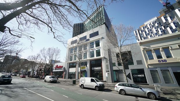 Street-level view of a commercial urban streetscape with a contemporary multi-storey office or retail building at the centre, featuring a grey masonry facade, large glazed windows, signage and a rooftop glass structure. Adjacent modern buildings, mature street trees, parked vehicles and marked roadway are visible.