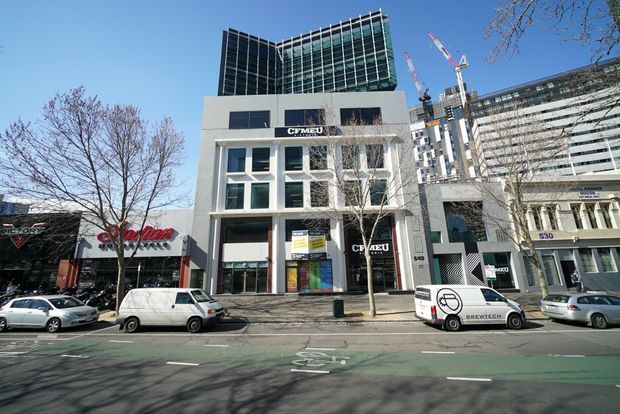 Street-level view of a contemporary multi-storey commercial building in an urban setting. The facade combines a light rendered lower section with extensive dark glazed curtain-wall office levels above, retail and commercial signage at ground level, and neighbouring commercial buildings and construction cranes visible along the street.