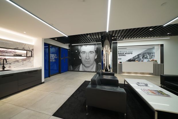 A contemporary commercial interior lobby or showroom reception area with a polished concrete-look tiled floor, dark built-in kitchenette or refreshment counter, black leather seating, a white reception desk, large graphic wall panels, and linear LED lighting. The ceiling combines smooth white panels with a black slatted feature section, creating a bold, modern corporate environment.
