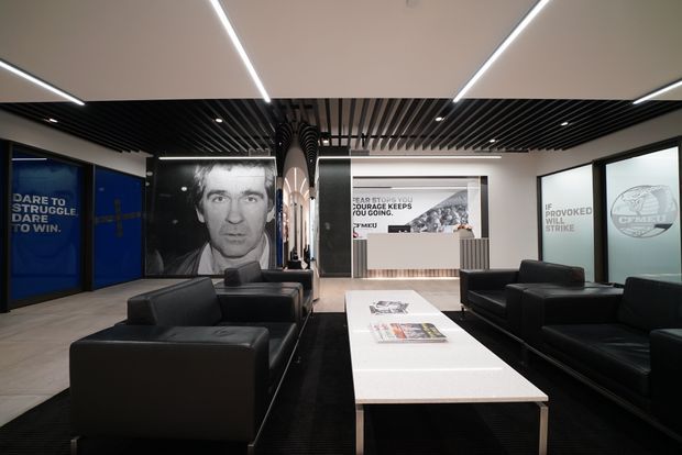 A contemporary commercial reception or waiting lounge with black leather sofas and armchairs arranged around a long white rectangular coffee table. The space features polished concrete-look flooring, white walls, black slatted ceiling panels, integrated linear LED lighting, dark-framed glazed partitions, and large monochrome graphic wall panels with bold motivational text and imagery. A reception counter or service desk is visible at the rear.