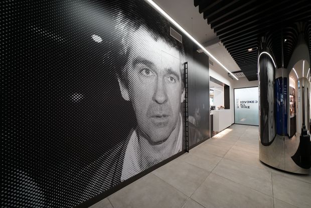 A sleek contemporary commercial interior corridor or reception area. A large black-and-white halftone portrait mural dominates the left wall, while reflective cylindrical and curved display elements line the right side. The space features large light-grey floor tiles, a linear LED ceiling strip, and a black slatted ceiling with integrated lighting. A glazed doorway and white reception or service area are visible at the end of the corridor.