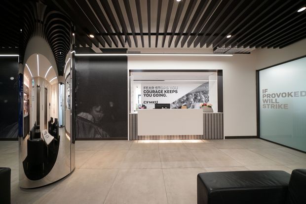 A contemporary commercial reception lobby with a pale tiled floor, white walls, black trim, and a central reception desk backed by a branded graphic. The space features a dramatic black slatted ceiling, curved black architectural screening and reflective surfaces on the left, recessed lighting, and black leather seating in the foreground. Glazed branding panels and a corridor are visible around the reception area.