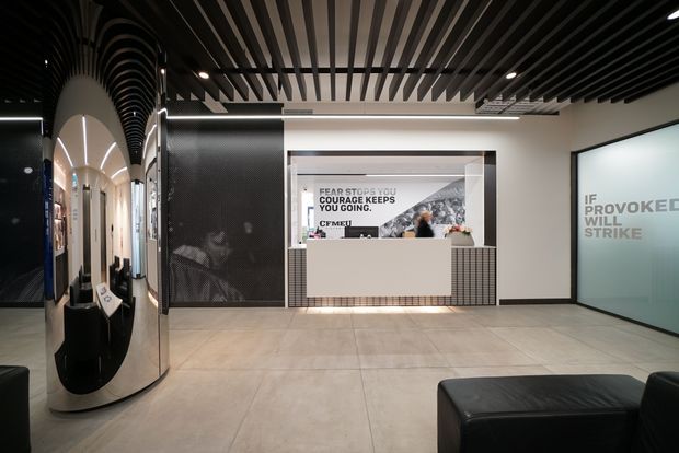 A contemporary commercial reception lobby with a dark, dramatic palette. The space features a curved reflective black feature wall with arched detailing, a central reception counter backed by a tiled wall and promotional display, large frosted glass lettering, pale large-format floor tiles, black seating, and a striking slatted ceiling with integrated linear lighting.