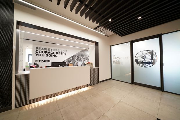 A modern commercial reception area with a pale built-in reception desk, tiled flooring, black-framed frosted-glass office doors, and a dramatic black slatted ceiling. Graphic wall and glazing decals feature union-themed typography and imagery, while integrated linear and recessed lighting create a refined, contemporary workplace entry.