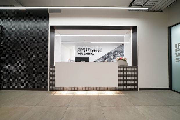 Modern commercial reception area with a clean white reception desk set within a recessed architectural opening. The space features black perforated and gridded wall panels, integrated linear lighting, a large graphic backdrop behind the desk, exposed office-style ceiling services, and large-format grey floor tiles.