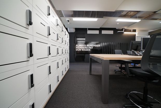 A contemporary commercial office interior with rows of white locker-style storage cabinets along the left wall, shared workstations and office chairs on the right, and a central rectangular meeting or work table. The space has dark carpet, black feature walls, exposed ceiling services, linear fluorescent lighting, and a motivational wall graphic.