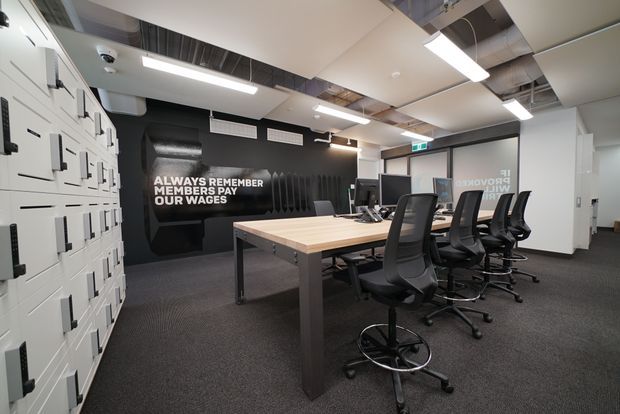 A contemporary commercial office or meeting space with a long light-wood worktable surrounded by black ergonomic chairs. The room features dark carpet, white walls and partitions, exposed ceiling services and ductwork, linear fluorescent-style lighting, black acoustic wall treatment with bold lettering, and extensive white built-in storage or lockers along the left wall. Glazed partitions and an illuminated exit sign are visible at the rear.