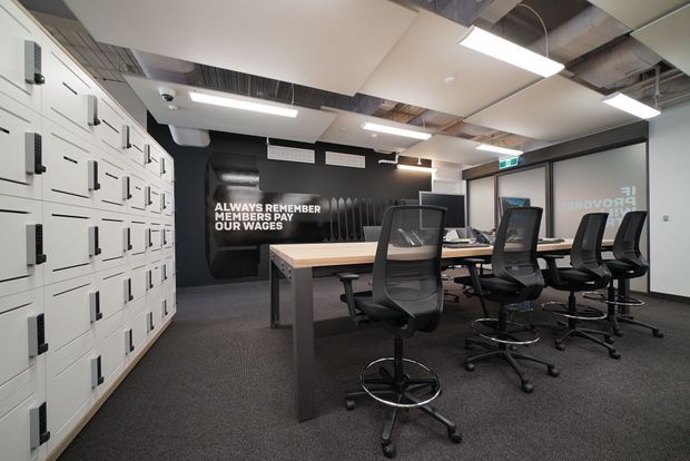 A contemporary commercial office or coworking space with a long shared workstation, multiple black ergonomic office chairs, extensive white locker-style storage along the left wall, dark acoustic wall treatment, and exposed ceiling services. Linear fluorescent and recessed lighting illuminate a neutral grey carpeted interior. A bold wall graphic reads “ALWAYS REMEMBER MEMBERS PAY OUR WAGES,” while glazed doors and an illuminated exit sign are visible at the rear.