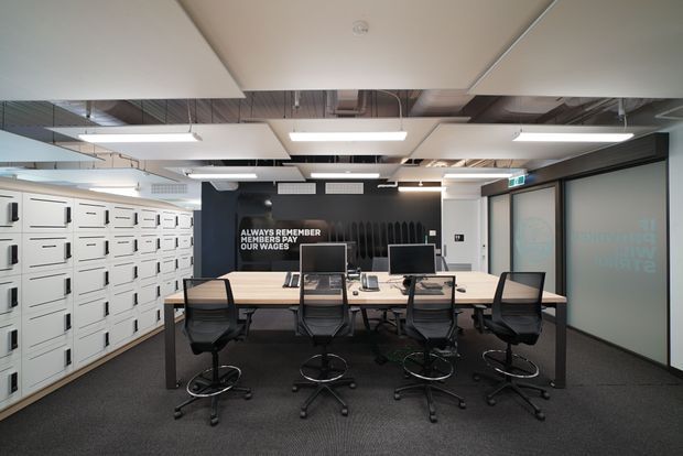 A contemporary commercial office workspace with a long shared meeting or hot-desk table, multiple black office chairs, computer monitors, and extensive white built-in locker storage along the left wall. The room has dark carpet, black and white finishes, exposed services in the ceiling, suspended linear lighting, acoustic ceiling panels, and glazed office doors.