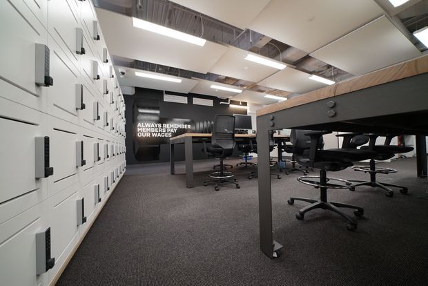 Open-plan commercial workspace with rows of large black desks and office chairs, a wall of white built-in storage lockers on the left, dark carpet flooring, and exposed services in the ceiling. The space has a utilitarian, contemporary office fit-out with linear fluorescent lighting and a motivational wall graphic.