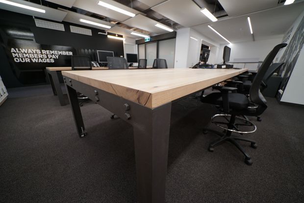 A contemporary open-plan commercial meeting or boardroom space dominated by a large rectangular light timber meeting table with dark metal legs. Black ergonomic office chairs surround the table, while black and white walls, exposed ceiling services, linear LED lighting, dark carpet, monitors and office equipment create a modern workplace setting. A motivational wall graphic is visible at the rear.