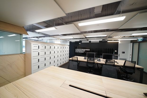Contemporary open-plan commercial office interior with a large pale timber communal worktable in the foreground, rows of desks and black office chairs behind it, extensive white built-in locker storage, and a black feature wall with white lettering. The space has exposed dark ceiling services, suspended linear fluorescent lighting, acoustic ceiling panels, glass partitions, and light timber wall cladding.