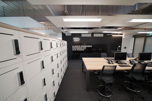 A contemporary commercial office or workplace interior with rows of white personal lockers along the left wall and a shared workstation table with computer monitors and ergonomic stools on the right. The space has an exposed services ceiling, linear fluorescent lighting, dark carpet, white cabinetry, and a black feature wall with motivational text. An exit door is visible at the rear.