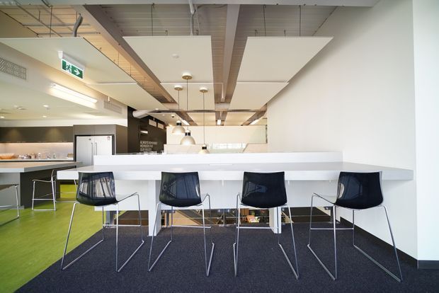Bright contemporary commercial interior featuring a long white counter or communal workbench with four black wire-frame chairs. The space has a mixed carpet floor in dark charcoal and vivid green, exposed ceiling services and beams, suspended acoustic ceiling panels, pendant lighting, and a kitchenette or service area in the background.