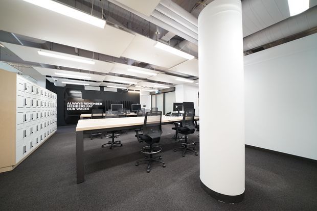 Open-plan commercial office interior with a long shared workstation, black mesh office chairs, computer monitors, extensive white locker storage along the left wall, dark carpet flooring, and exposed ceiling services. A large white structural column dominates the foreground, while linear fluorescent-style lighting and visible ductwork contribute to an industrial office fit-out. A black feature wall at the rear carries white lettering.