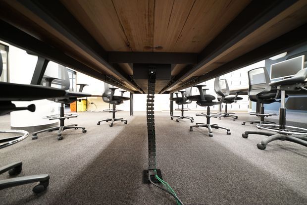 Low-angle view beneath a long shared office workstation, showing exposed dark structural beams, a timber underside, cable management, carpeted flooring, and rows of ergonomic rolling office chairs in a bright open-plan workspace.