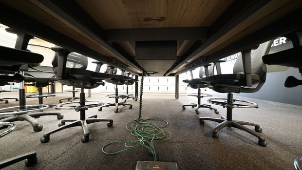 Low-angle view beneath long office workstations, showing rows of ergonomic wheeled office chairs, exposed desk supports, cable management, grey carpet flooring, and a timber-lined ceiling. The image appears to document a commercial office fit-out or workspace installation.