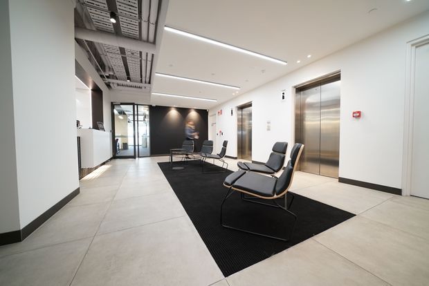 A contemporary commercial lift lobby or reception waiting area with two stainless-steel elevator doors, minimalist black lounge chairs arranged on a large black rug, pale tiled flooring, white walls, linear recessed lighting, and an exposed services ceiling section. A dark feature wall and glazed entry or corridor appear at the rear.