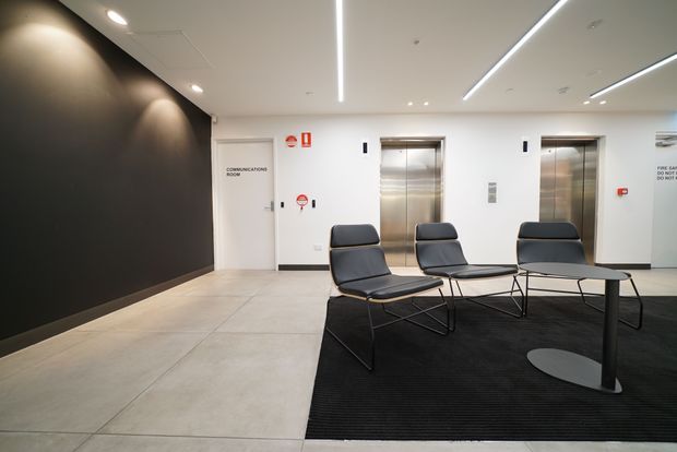 A contemporary commercial lobby or lift waiting area with two stainless-steel elevator doors, a white ceiling with linear recessed LED lighting, a dark feature wall, pale large-format floor tiles, and black lounge chairs arranged around a small table on a black rug. Wayfinding and safety signage are visible on the walls.