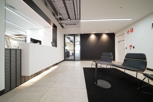Contemporary commercial reception and waiting area with a minimalist white reception desk, black feature wall, glass entry doors, exposed ceiling services, linear lighting, and a black lounge seating arrangement over a dark rug. Large-format pale flooring and strong black-and-white contrast create a clean, refined professional atmosphere.