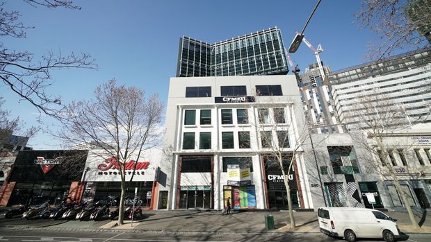 Street-level exterior photograph of a contemporary multi-storey commercial building in an urban streetscape. The white facade has large glazed windows, dark rectangular panels, ground-floor retail or showroom entrances, and a prominent curved glass upper level. Adjacent commercial buildings, signage, parked motorcycles, a van, trees, and a streetlight are visible.