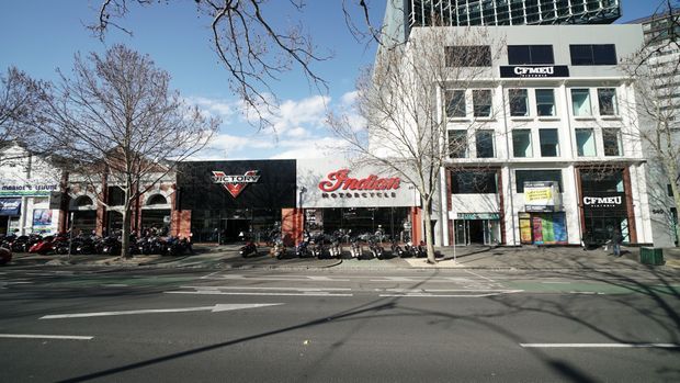 Street-level view of a commercial urban streetscape with a row of retail and business buildings, including prominent Victory and Indian motorcycle dealerships. The scene shows multi-storey shopfronts, large display windows, signage, parked motorcycles, mature street trees, and a broad road in the foreground.