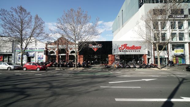 Street-level exterior view of a contemporary multi-storey commercial building with ground-floor motorcycle dealerships and retail storefronts. The facade combines large glazed sections, white rendered surfaces, dark shopfront signage, and upper-level windows. A broad paved roadway and sidewalk with leafless street trees occupy the foreground.