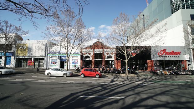 Street-level view of a contemporary urban commercial shopping strip with several adjoining retail storefronts, prominent signage, large glazed shopfronts, and a taller light-coloured building volume above. Leafless street trees, parked cars, bicycles, and a broad paved roadway are visible in the foreground.