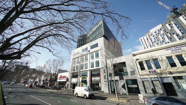 Street-level view of a dense urban commercial streetscape with a modern multi-storey office or mixed-use building in the centre, featuring a glass-and-metal upper volume, white facade, large windows, and ground-floor retail or business signage. Adjacent older commercial buildings, mature street trees, parked vehicles, and construction activity are visible.